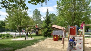 A playground with various play structures, including a swing set and climbing frames, surrounded by trees and a sandy area. - Camping Le Petit Lion - Gites (Vendoire)