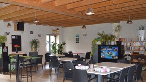 A café with multiple tables and chairs, wooden ceiling beams, and a television screen displaying a program. - Camping Le Petit Lion - Gites (Vendoire)