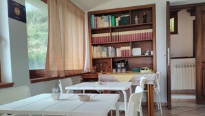 Une bibliothèque avec des tables et des chaises blanches, une étagère remplie de livres et une fenêtre avec des rideaux.