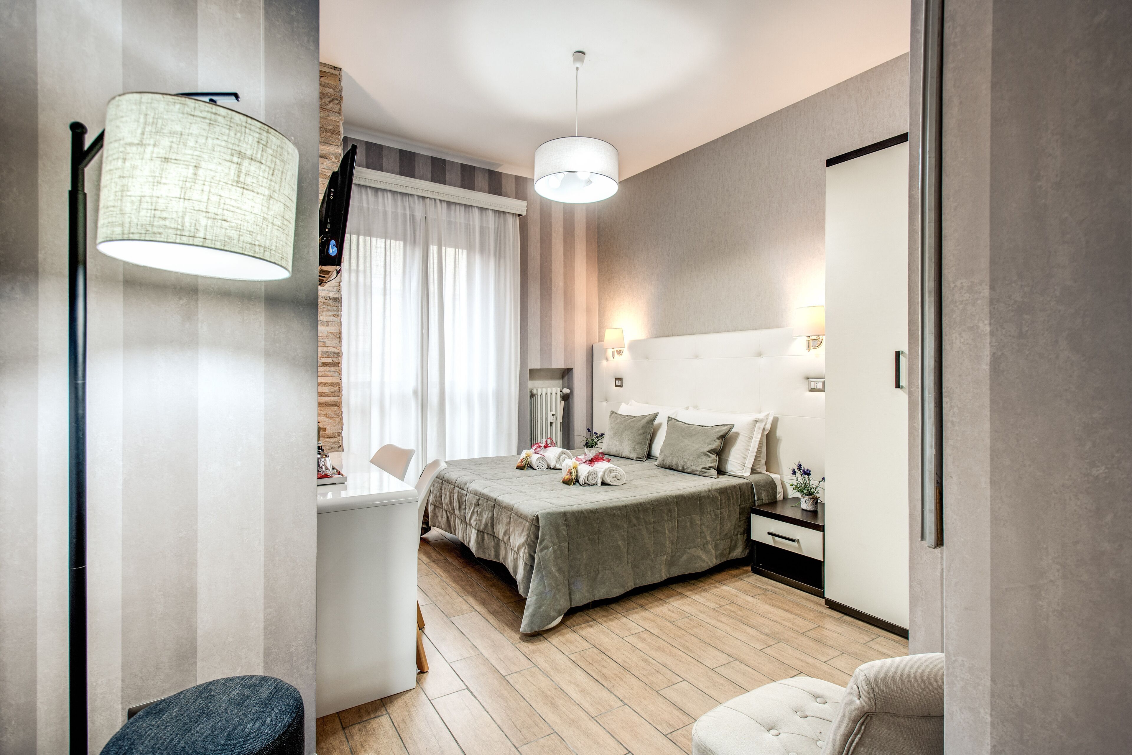 A modern hotel room with a bed, bedside table, lamp, and a desk with a chair.