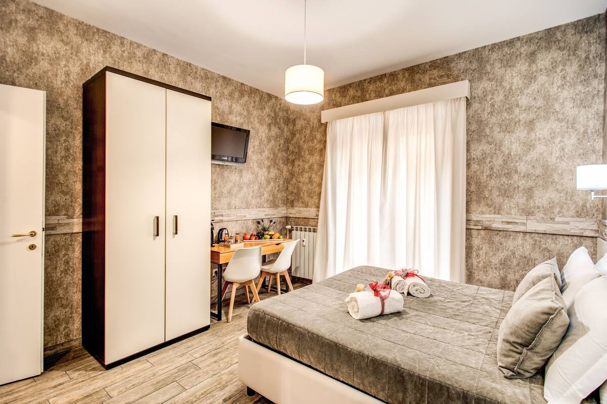 A hotel room with a bed, a desk with a chair, a wardrobe, and a television on the wall.