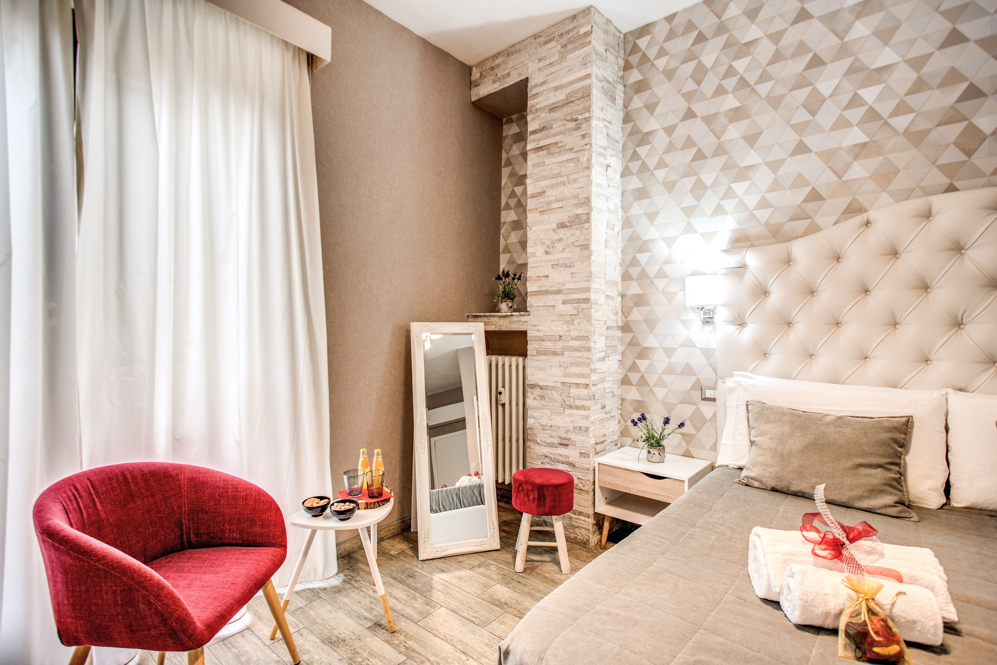 Une chambre d’hôtel avec un fauteuil rouge, un lit doté d’une tête de lit capitonnée, une petite table avec des articles de toilette et un miroir.