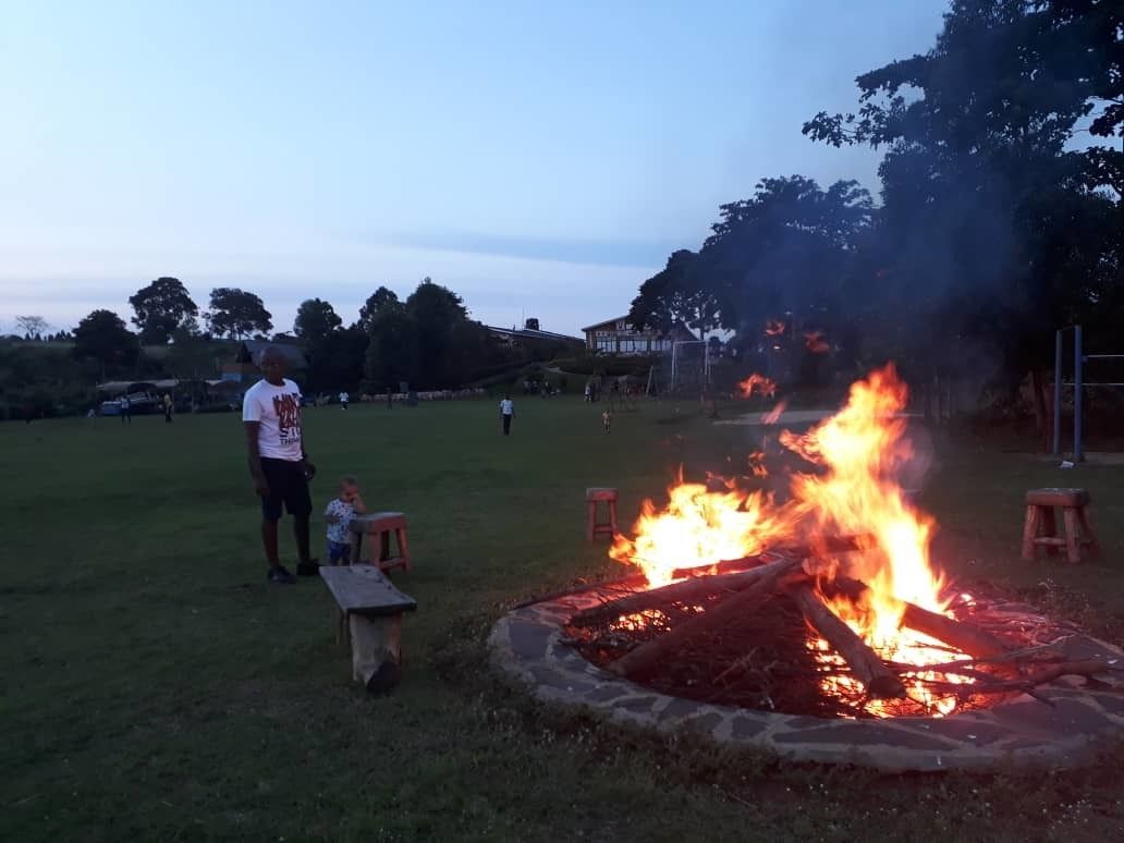 Una fogata en un campo cubierto de hierba, con un pozo de piedra, rodeado de árboles y algunas personas al fondo.