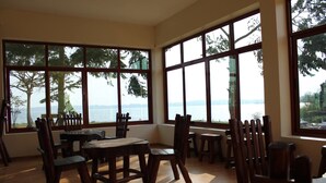 A dining area with large windows overlooking a body of water, wooden furniture, and a view of trees and hills. - Nyange Resort and Marina (Entebbe)