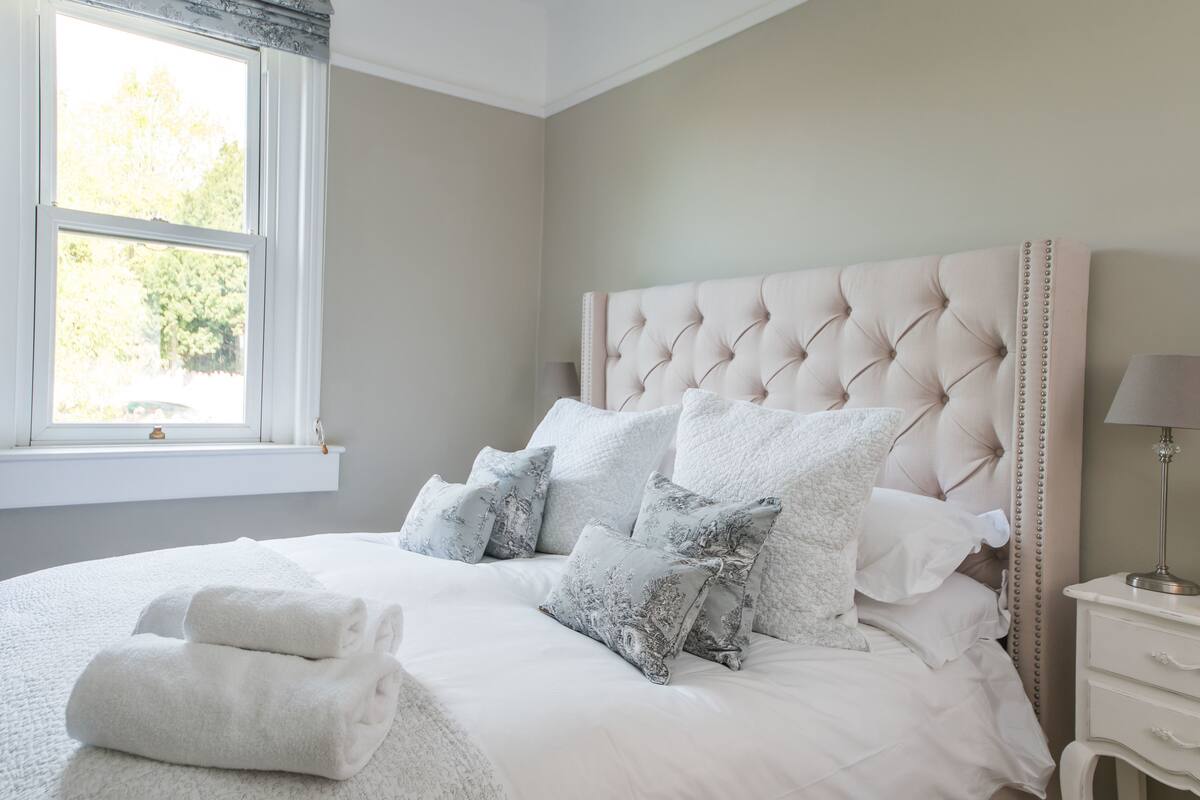 Un lit bien fait, avec du linge de lit blanc et des coussins décoratifs, une lampe de chevet et une fenêtre donnant sur des arbres.