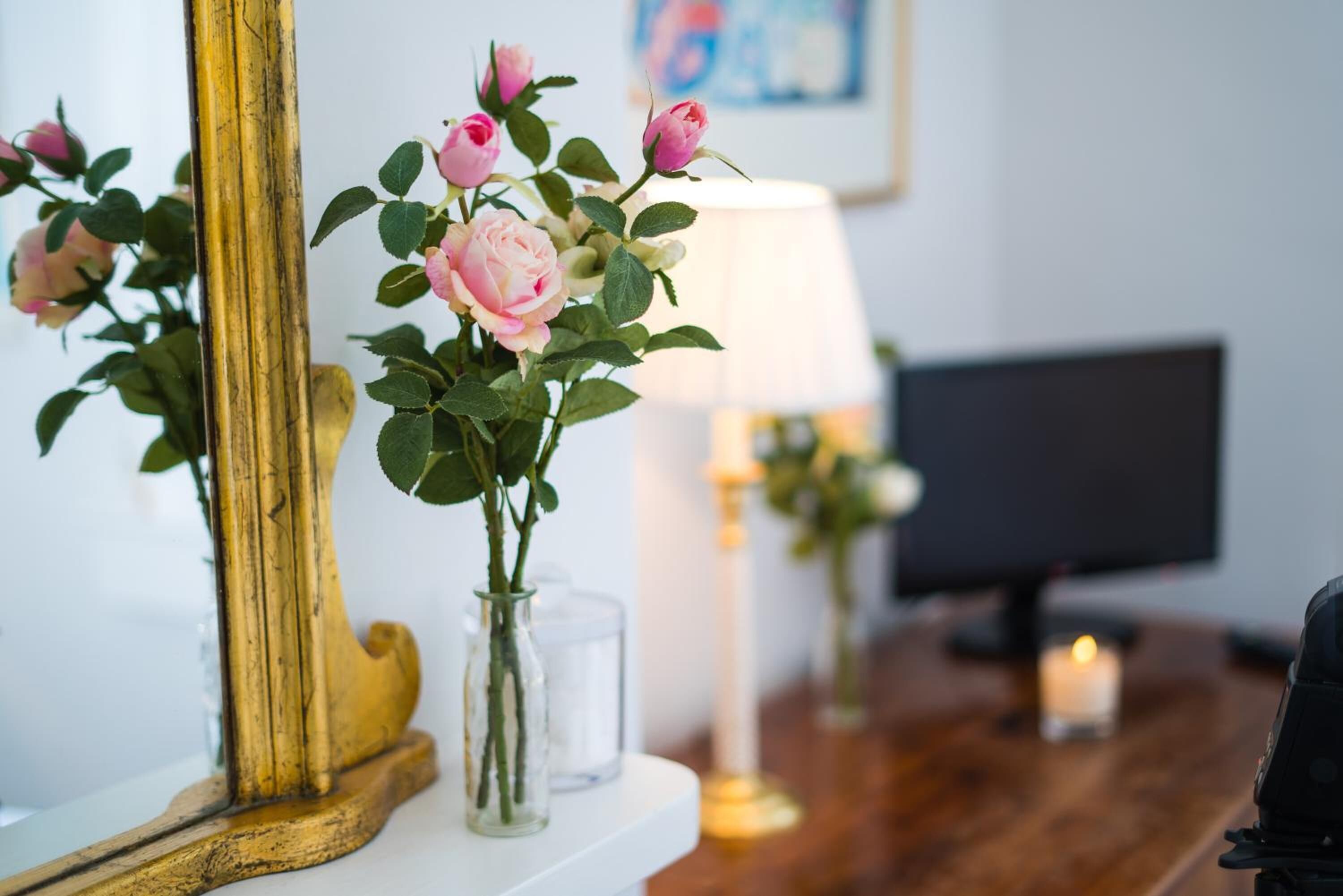 A room with a mirror, a vase with flowers, a lamp, and a computer monitor.