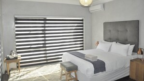 Een slaapkamer met een groot bed, een nachtkastje, een bankje, een hangende lamp en een raam met jaloezieën.