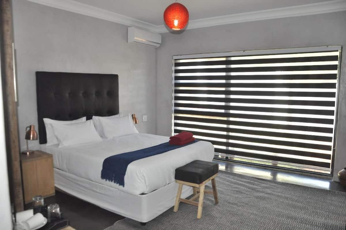 Une chambre d’hôtel moderne avec un grand lit, une table de chevet, une lampe rouge et une fenêtre avec des stores horizontaux.