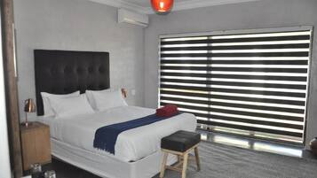 A modern hotel room with a large bed, a bedside table, a red lamp, and a window with horizontal blinds.