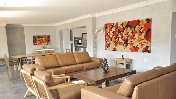 Een moderne woonkamer met een bruine leren bank, houten stoelen, een salontafel en een groot abstract schilderij aan de muur.