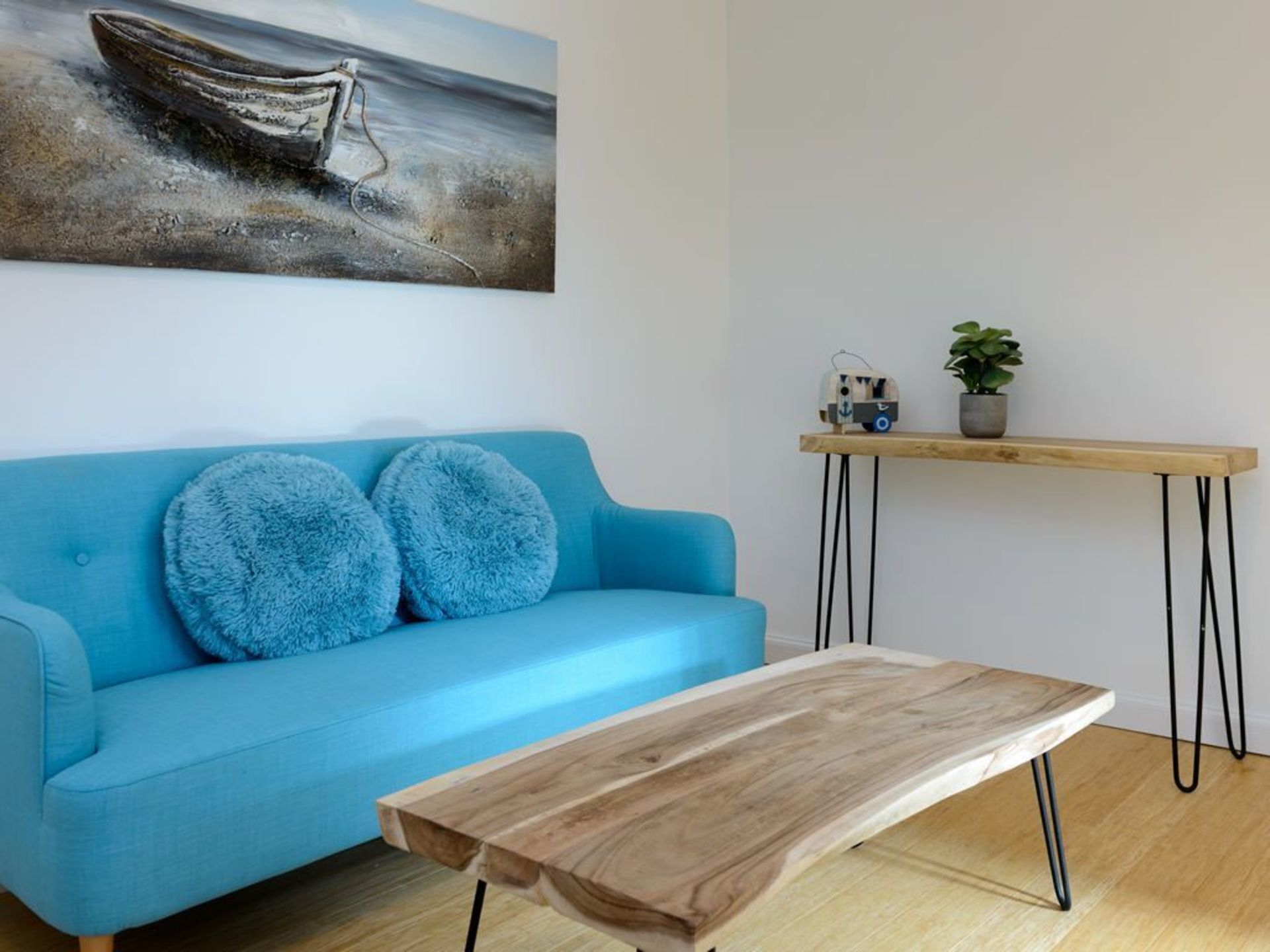 A room with a blue sofa, two fluffy pillows, a wooden coffee table, a wooden side table with a plant, and a painting of a boat on the wall.