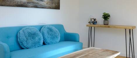 A room with a blue sofa, two fluffy pillows, a wooden coffee table, a wooden side table with a plant, and a painting of a boat on the wall.