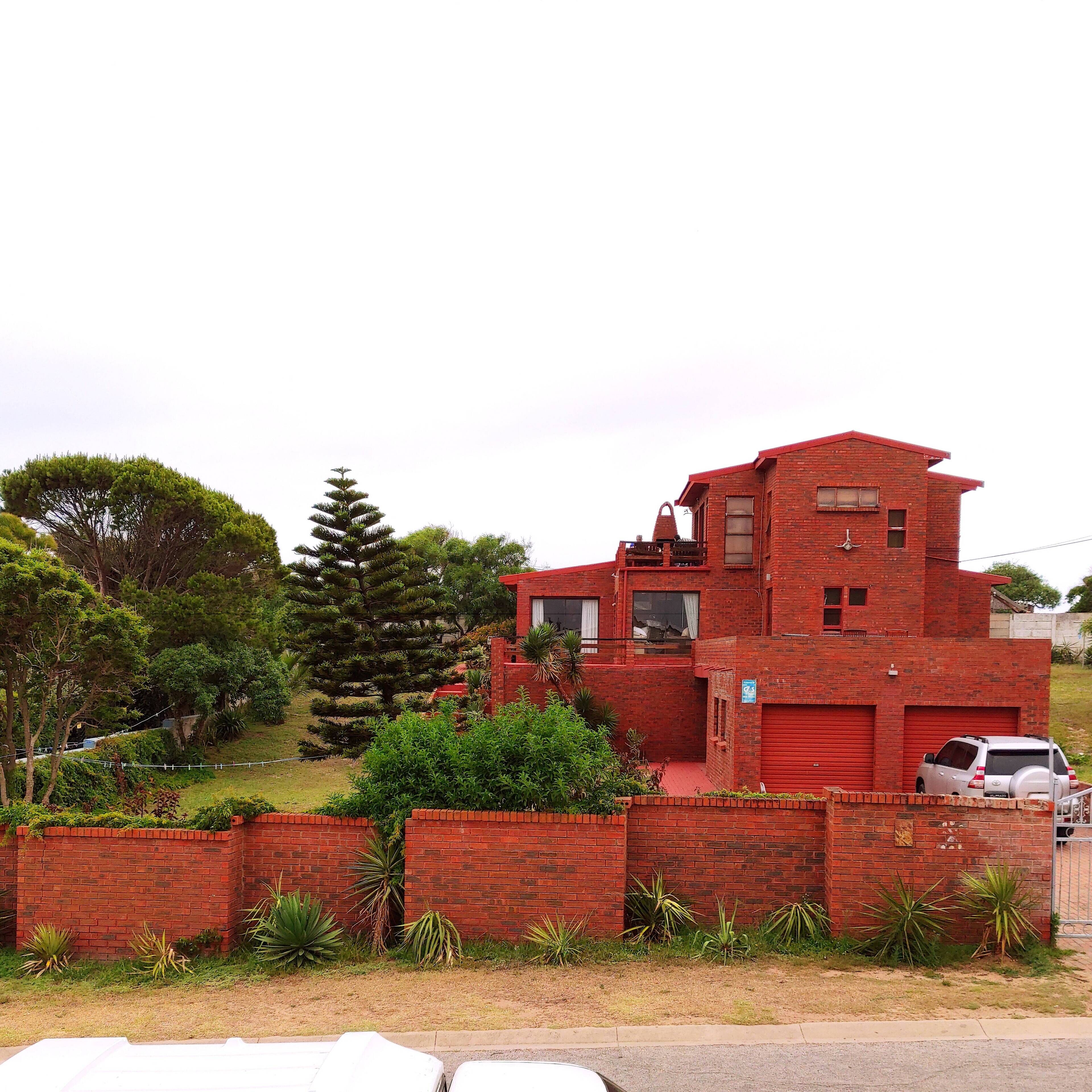 A red brick house with a small balcony, surrounded by greenery and a parked car.