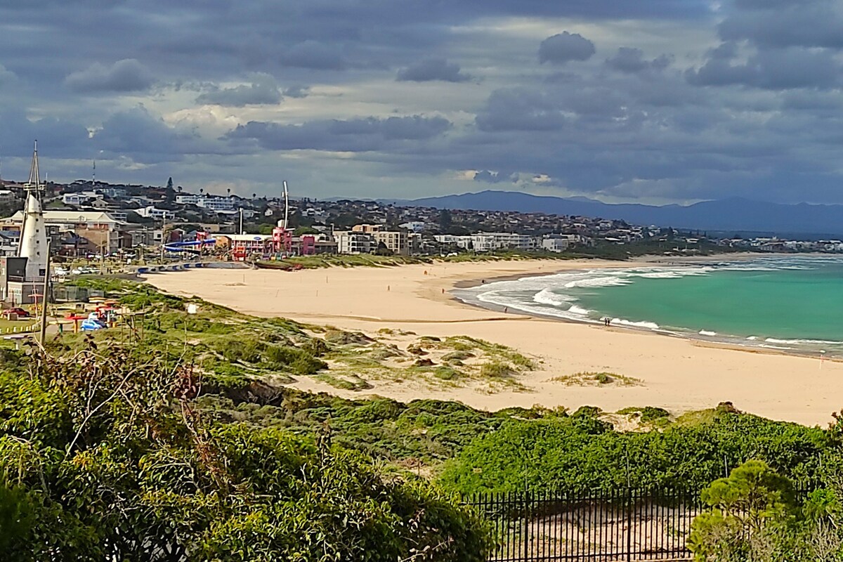 Un paysage côtier avec une plage de sable, une vue dégagée sur l’océan et une silhouette de ville visible au loin.