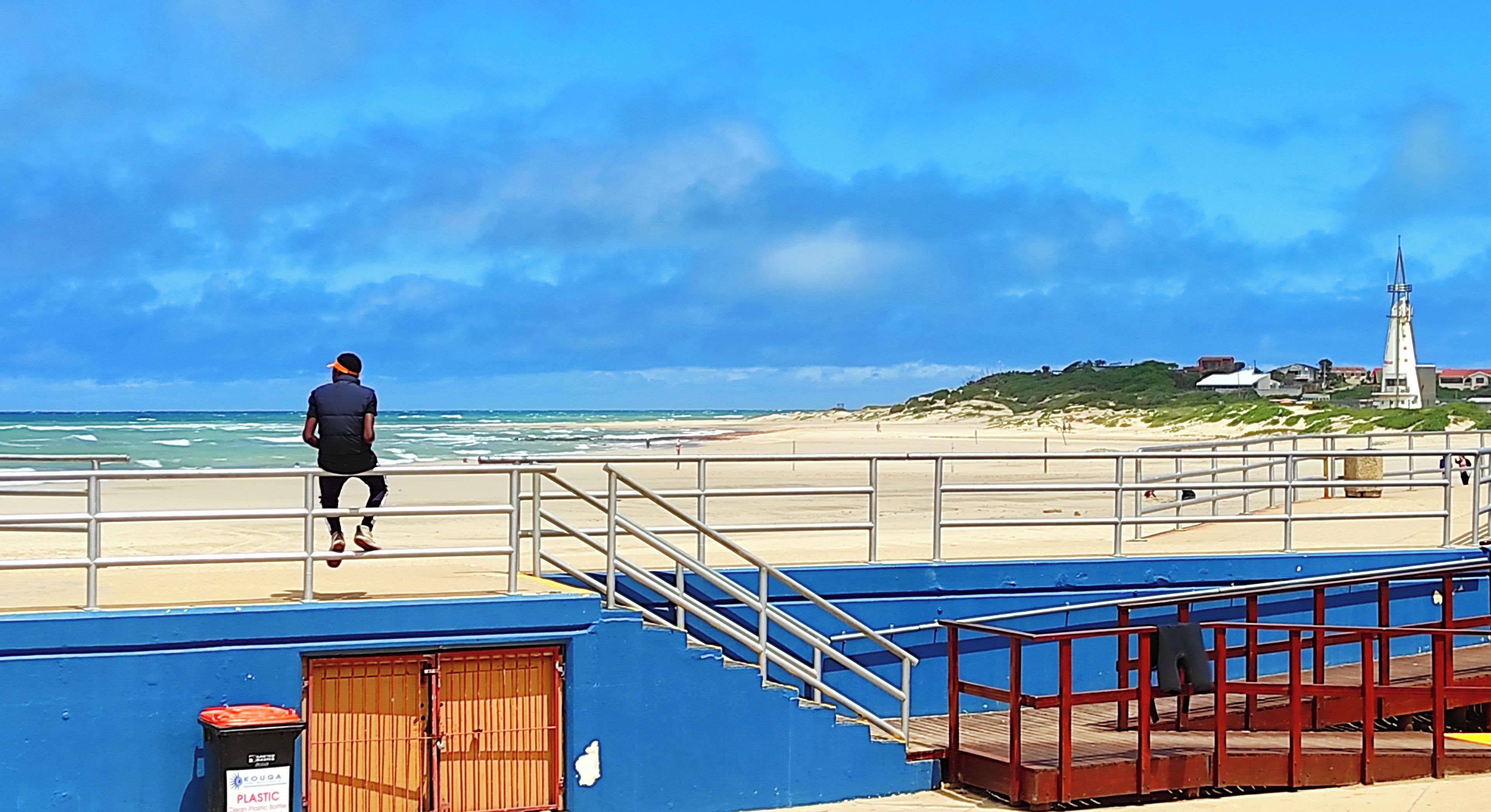 A coastal scene with a person walking along a blue railing, a white building with a pointed roof, and a beach in the background.