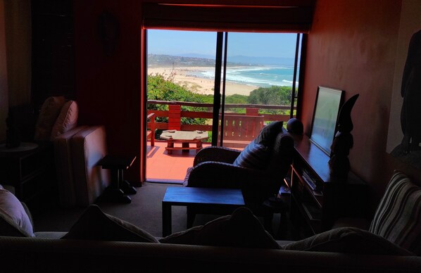 A living room with a large sliding glass door leading to a balcony with a view of the beach and ocean.