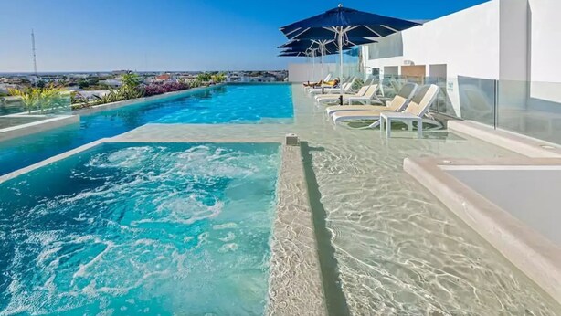 Une piscine à débordement située sur le toit, avec une eau d’un bleu cristallin, entourée de chaises longues et de parasols, offrant une vue imprenable sur le paysage urbain.