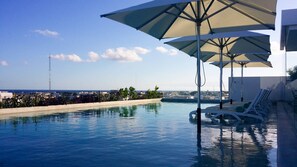 Una piscina con sillones de descanso y sombrillas, rodeada por un muro bajo y plantas, con vista a la ciudad y al océano al fondo.