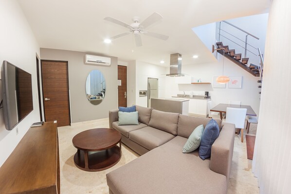 A modern living room with a sofa, coffee table, and a TV. There is a staircase leading to an upper level.