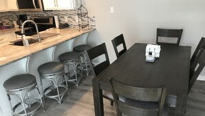 Un comedor con una mesa de madera y taburetes de barra metálicos, una cocina con microondas y pileta, y un salpicadero de azulejos.