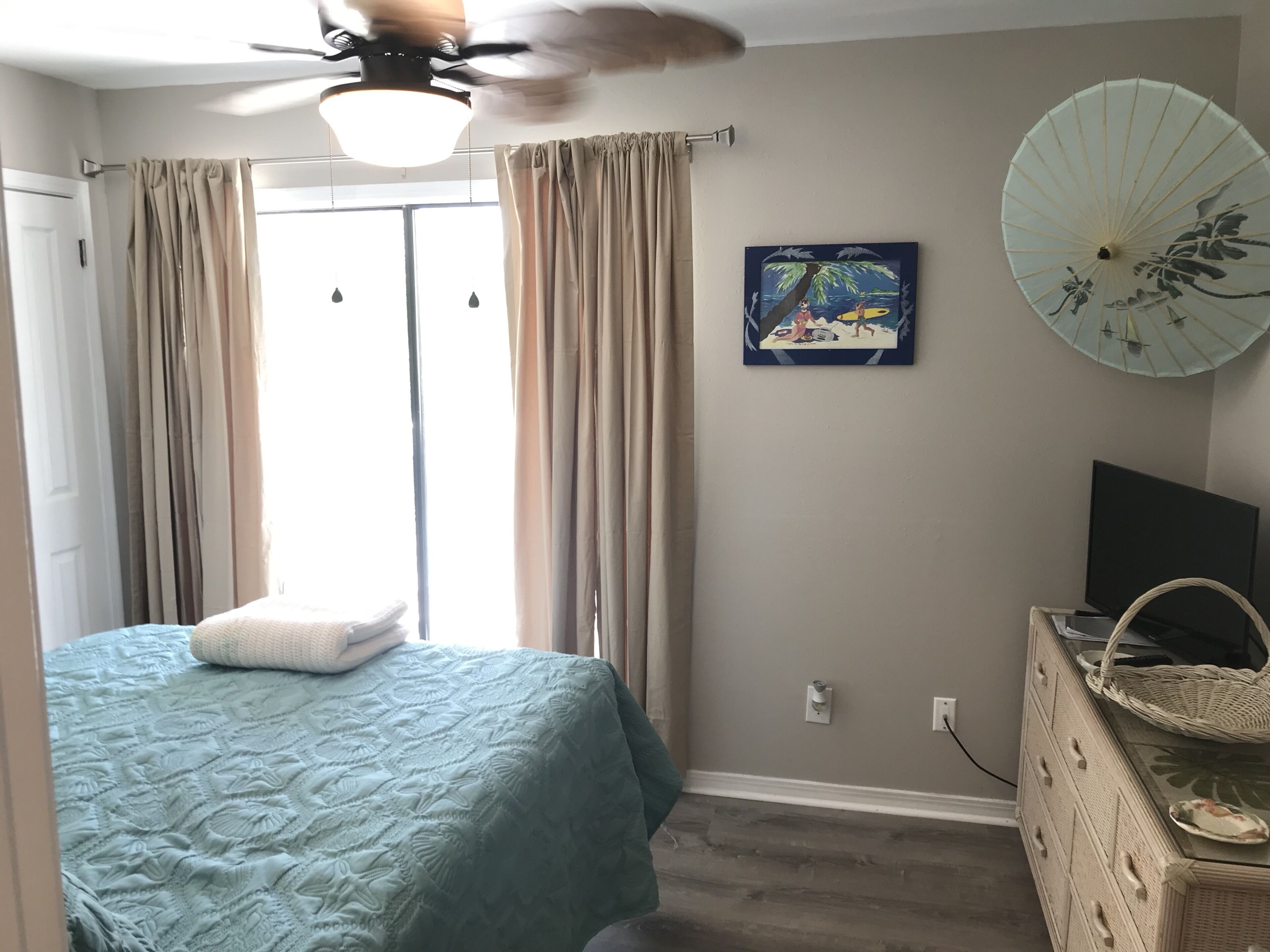 Un dormitorio con cama, ventilador de techo, ventana con cortinas, televisor montado en la pared y un ventilador decorativo.
