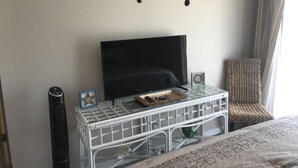 Un dormitorio con televisión sobre un mueble blanco, una silla de mimbre y una cama con manta estampada.