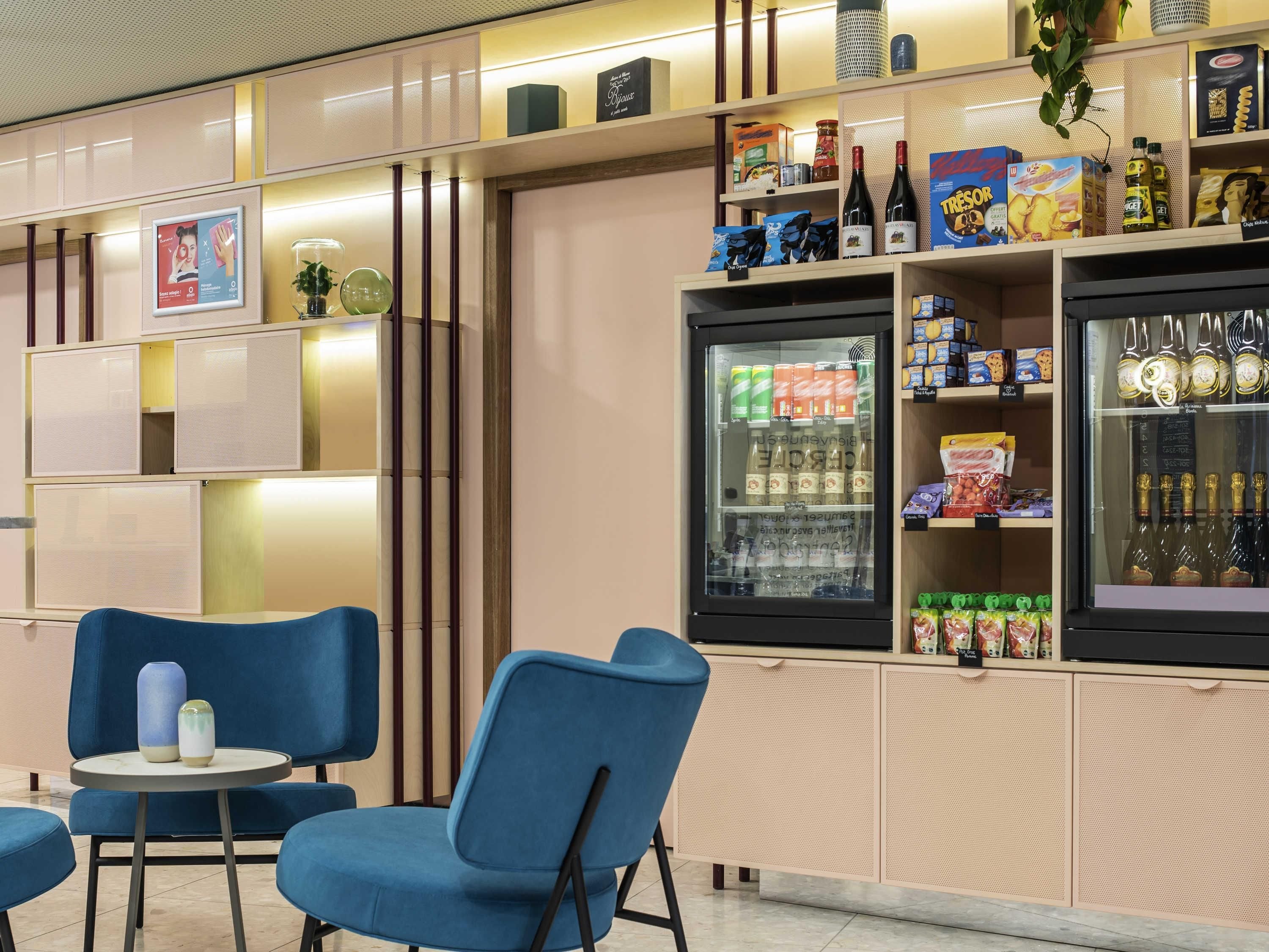 Una zona de estar con sillas azules y una mesita, un refrigerador con bebidas y estantes llenos de aperitivos y aderezos.