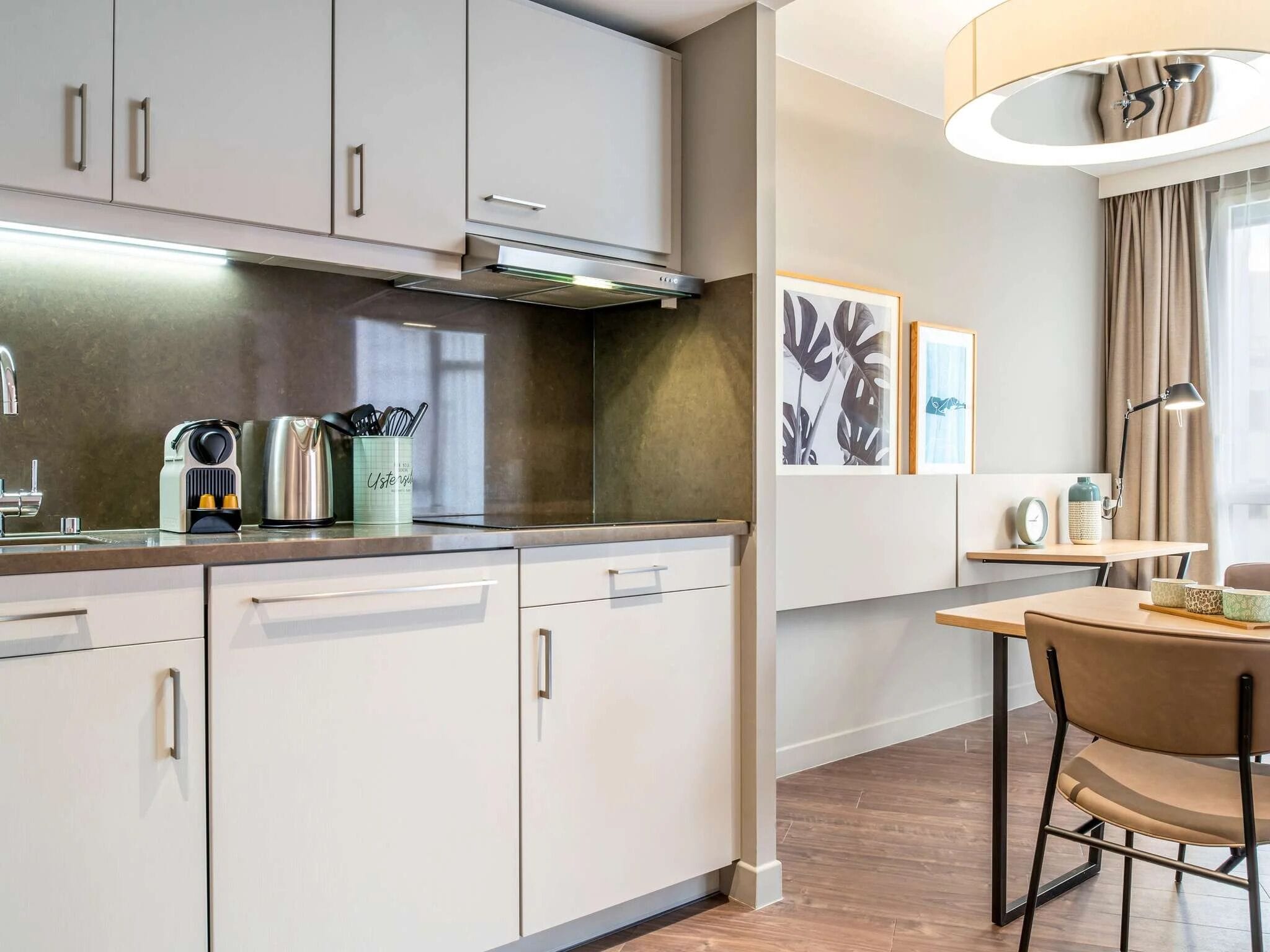 Una cocina moderna con armarios blancos, un hervidor de acero inoxidable y una cafetera. Hay una zona de comedor con una mesa y sillas, y una lámpara de pared.