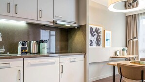 Una cocina moderna con armarios blancos, un hervidor de acero inoxidable y una cafetera. Hay una zona de comedor con una mesa y sillas, y una lámpara de pared.