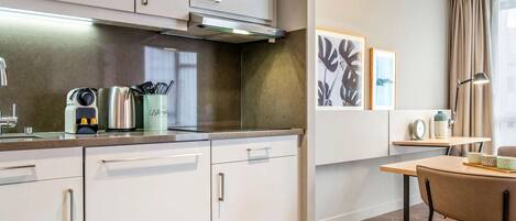 A modern kitchen with white cabinets, a stainless steel kettle, and a coffee maker. There is a dining area with a table and chairs, and a wall-mounted lamp.