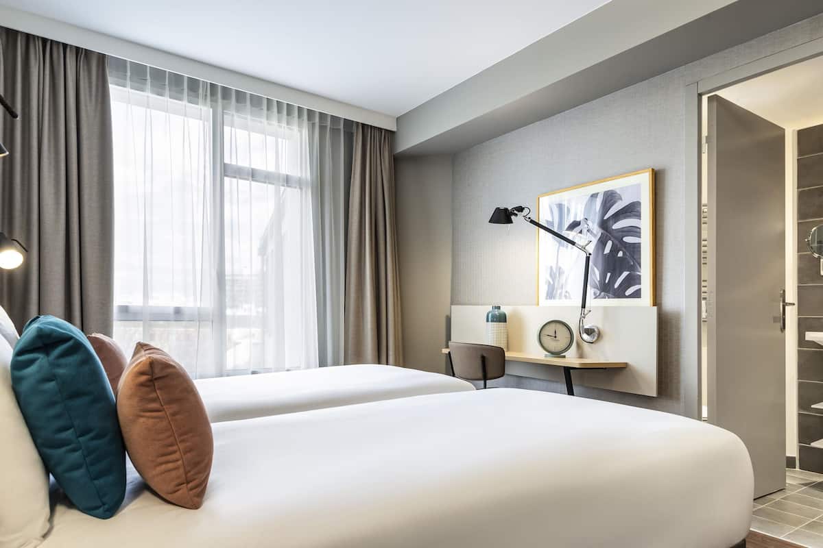 A hotel room with two beds, a desk with a lamp, a clock, and a framed picture on the wall.