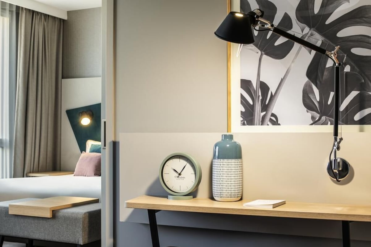 Habitación de hotel con cama, escritorio con lámpara, un reloj y una botella de agua.