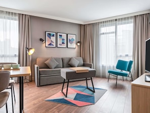 Una habitación de hotel moderna con sofá, un sillón azul, televisor, escritorio y mesa de centro.