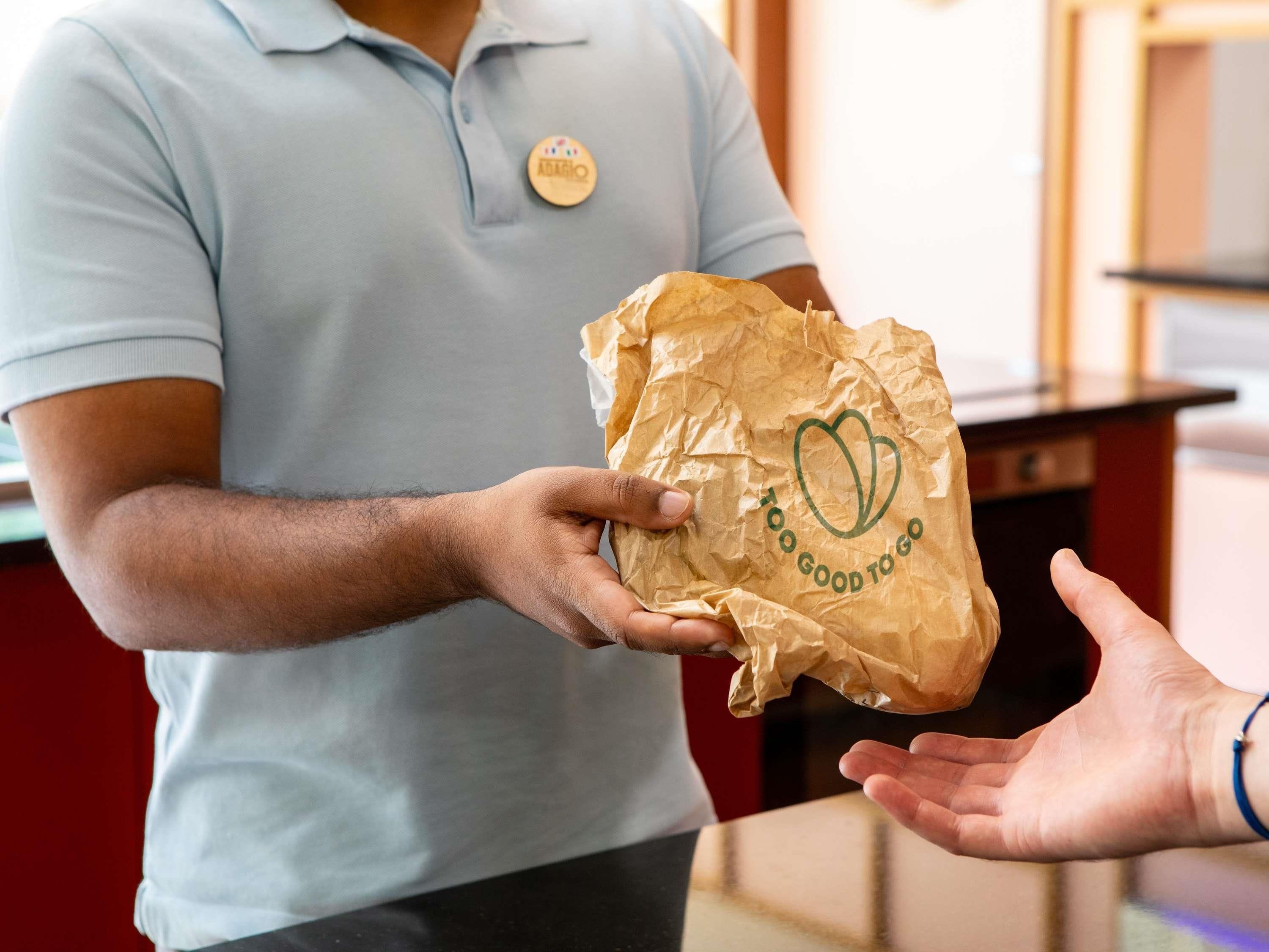 A person in a light blue shirt handing a crumpled brown paper bag with a logo to another person.