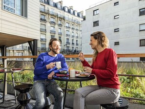Deux personnes sont assises à une table à l’extérieur, avec une vue sur le paysage urbain. On aperçoit une chaise en métal et une petite table sur laquelle se trouvent une tasse et une assiette.