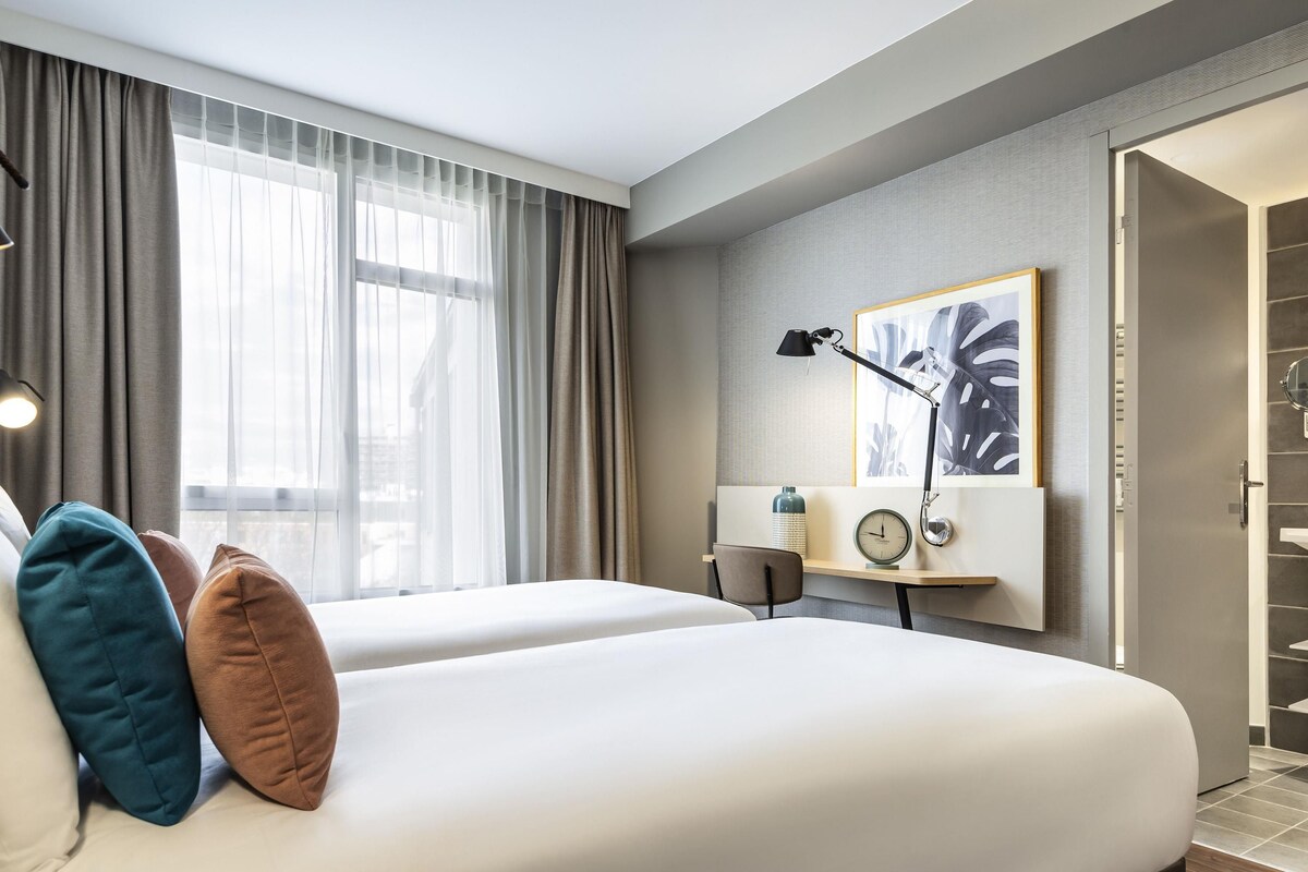 A hotel room with two beds, a desk with a lamp, a clock, and a framed picture on the wall.