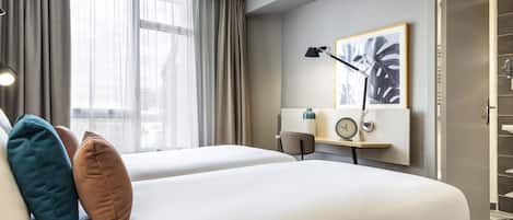 A hotel room with two beds, a desk with a lamp, a clock, and a framed picture on the wall.