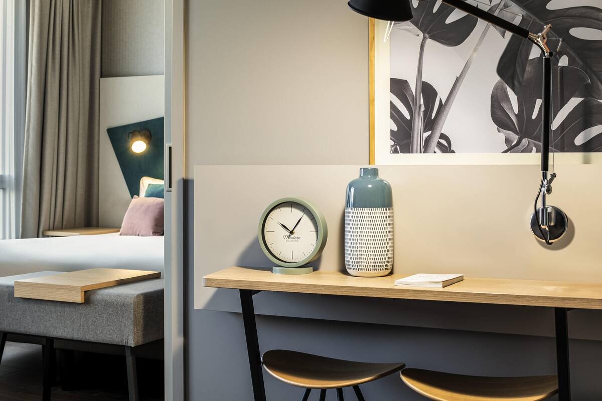 A modern hotel room with a bed, a desk with a lamp, a clock, and a decorative vase.