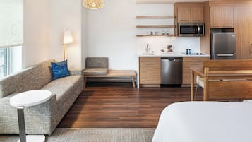 A modern, compact hotel room with a sofa, a small table, a bed, a kitchenette, and a view of the city.