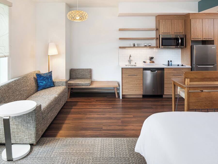 A modern, compact hotel room with a sofa, a small table, a bed, a kitchenette, and a view of the city.