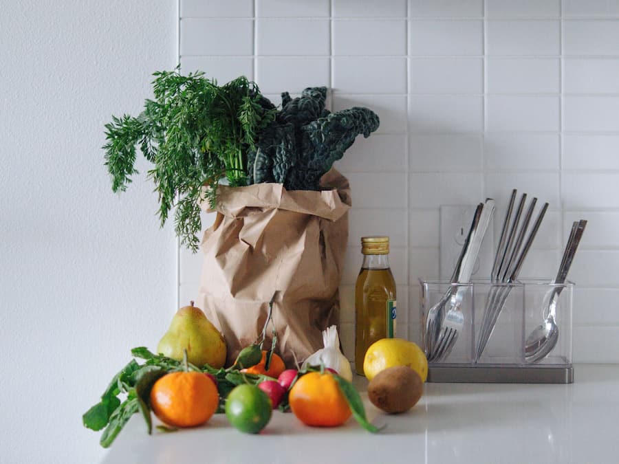 Una encimera de cocina con productos frescos, una bolsa de papel, una botella de aceite y utensilios.