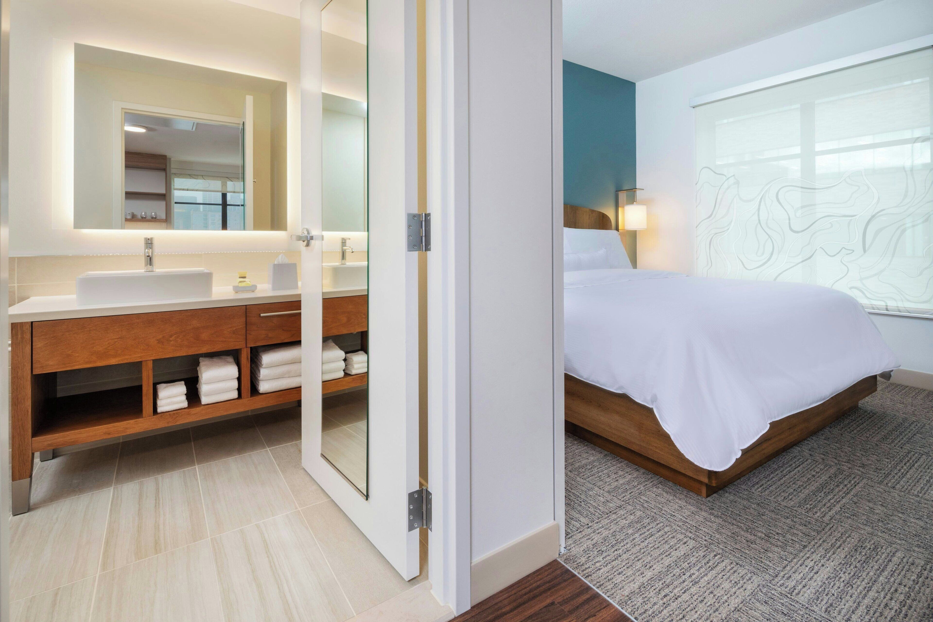 A modern hotel room with a large mirror, wooden vanity, white linens, and a large window with a view.