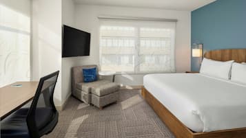 A hotel room with a bed, a desk with a chair, a small ottoman, a TV mounted on the wall, and a window with a view.