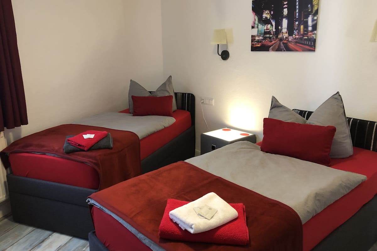 A hotel room with two beds, each with red and grey bedding, a nightstand with a lamp, and a cityscape painting on the wall.