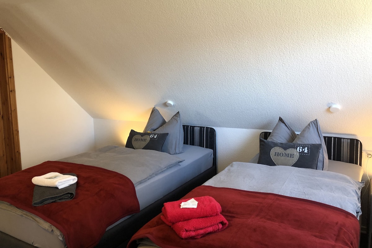 A bedroom with two beds, each with a grey headboard and pillows, and a red blanket on one bed.