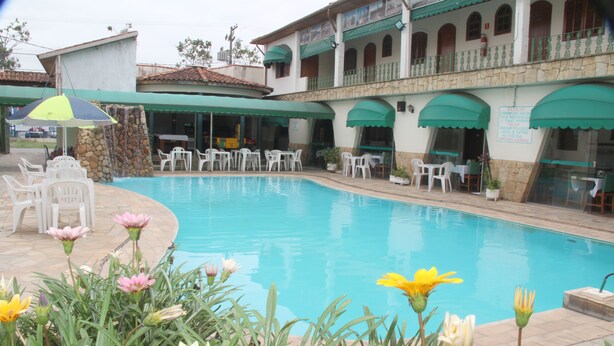 A swimming pool with surrounding white chairs and tables, a stone wall, and a building with green awnings. - Hotel Chalé São Jorge (Aparecida)