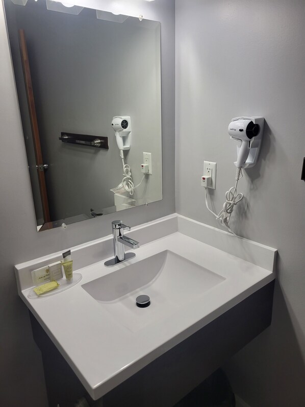 A bathroom with a white sink, a mirror, and two wall-mounted hair dryers. - Grenfell Heritage Hotel & Suites (St. Anthony)