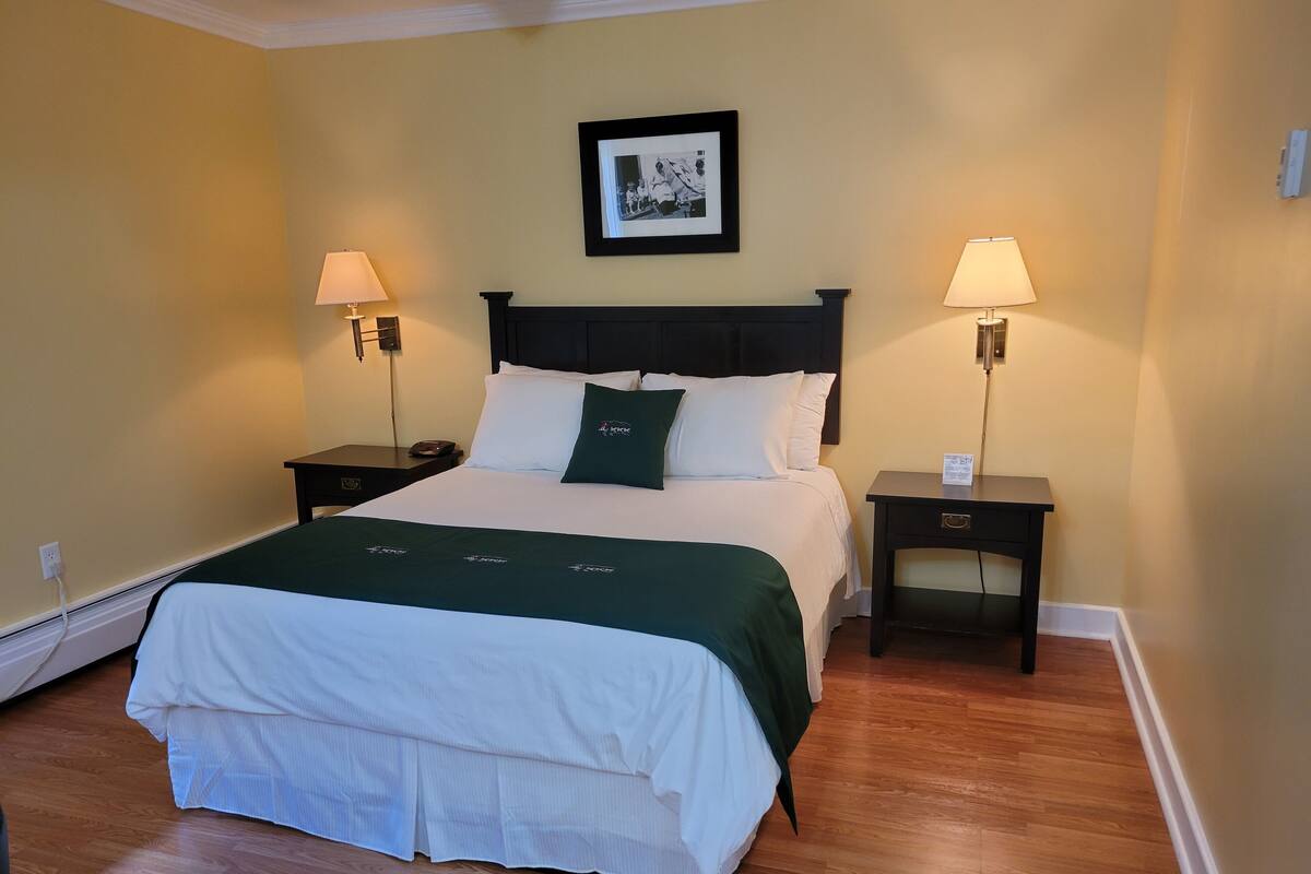 Een net opgemaakt bed met een groene deken, twee nachtkastjes met lampen, een ingelijste foto boven het bed, en een houten vloer.