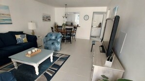 Een moderne woonkamer met een bank, salontafel en televisie. Er is een eethoek met stoelen en een tafel. In de hoek staat een grote plant.