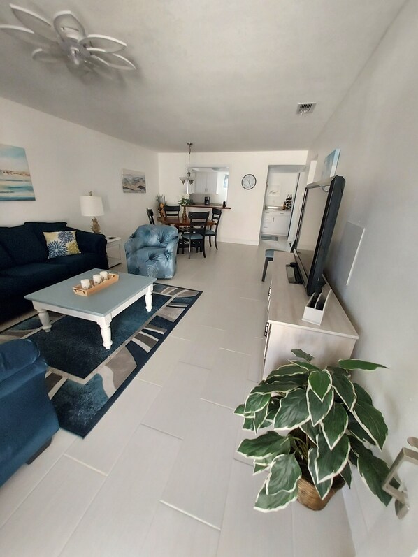 A modern living room with a sofa, coffee table, and a TV. There is a dining area with chairs and a table. A large plant is visible in the corner.